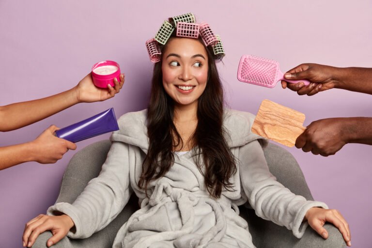 Image of attractive brunette Asian lady looks aside with smile, wears hair curlers, prepares for special occasion, wants to look alluring, gets grooming from beautician, isolated on purple wall