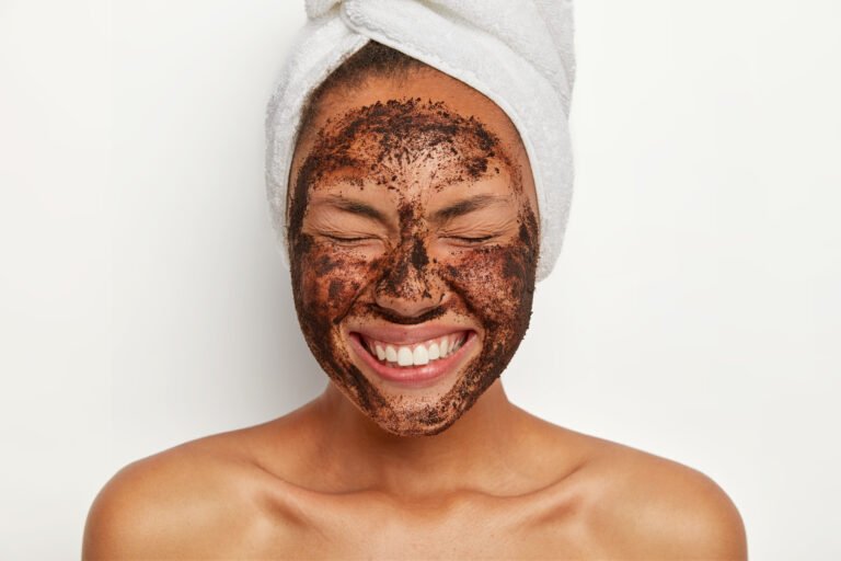 Headshot of good looking overjoyed young woman keeps eyes shut, smiles broadly, shows white perfect teeth, wears wrapped towel on head, peels face with coffee scrub, stands shirtless indoor